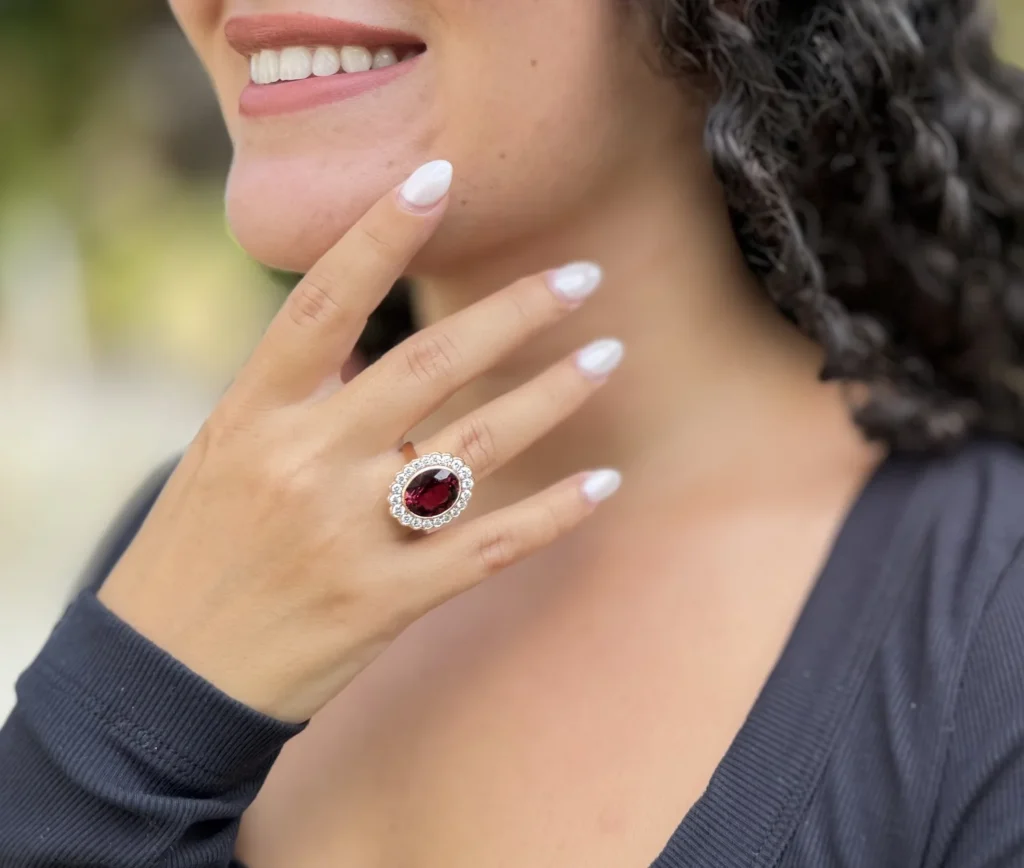 Close-up of a smiling woman wearing a large oval crimson gemstone ring surrounded by small diamonds, with white manicured nails and a soft blurred outdoor background.