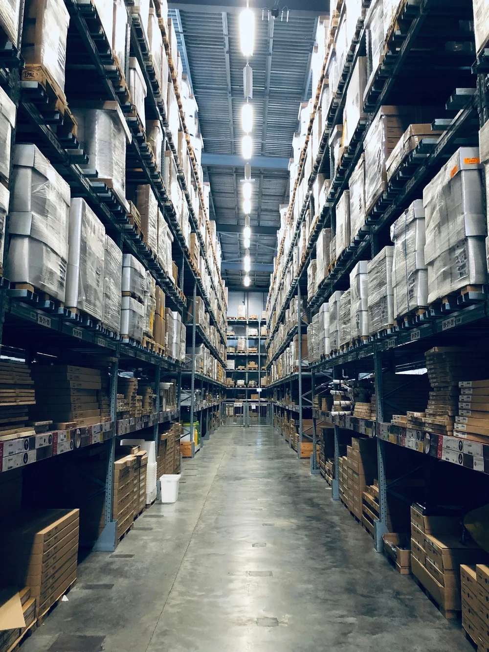 Long aisle inside a large warehouse with tall industrial shelving units filled with stacked boxes and wrapped pallets under bright overhead lights.