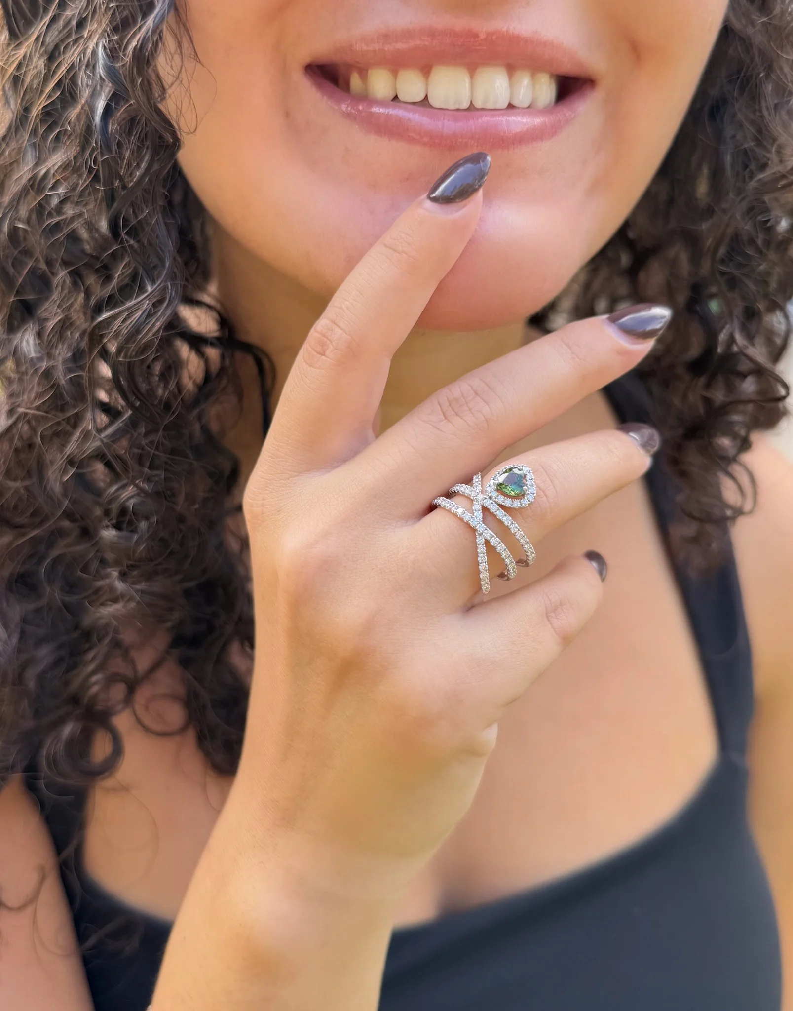 Close-up of a smiling woman with curly hair wearing a silver multi-band ring with a green teardrop gemstone and dark gray nail polish.